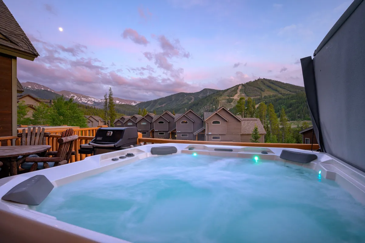 A private deck featuring a bubbling hot tub with unobstructed mountain views, an outdoor seating area, and a barbecue grill. The setting offers a tranquil ambiance, with the sun setting behind the peaks and space to relax or socialize with friends.