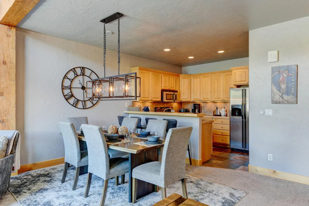 A dining space with a large wooden dining table illuminated by a chandelier, surrounded by kitchen cabinetry and subtle decor. The open floor plan connects the dining area seamlessly with the kitchen, creating a welcoming spot for shared meals and celebrations.