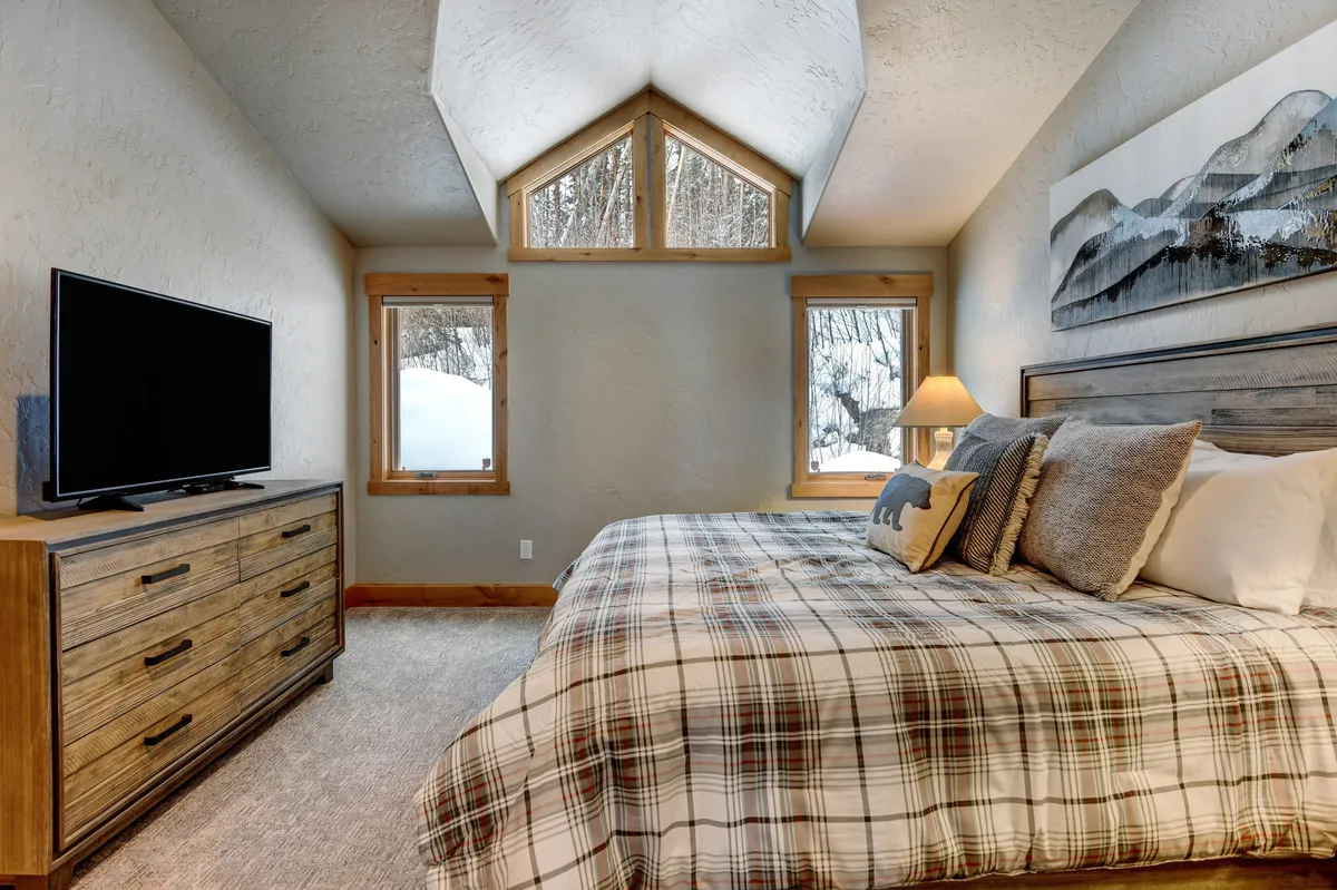 A cozy and serene bedroom with a bed dressed in plaid bedding, a wooden dresser with a TV, large windows allowing in natural light, and mountain-themed artwork. The setting radiates comfort and tranquility, perfect for restful nights.
