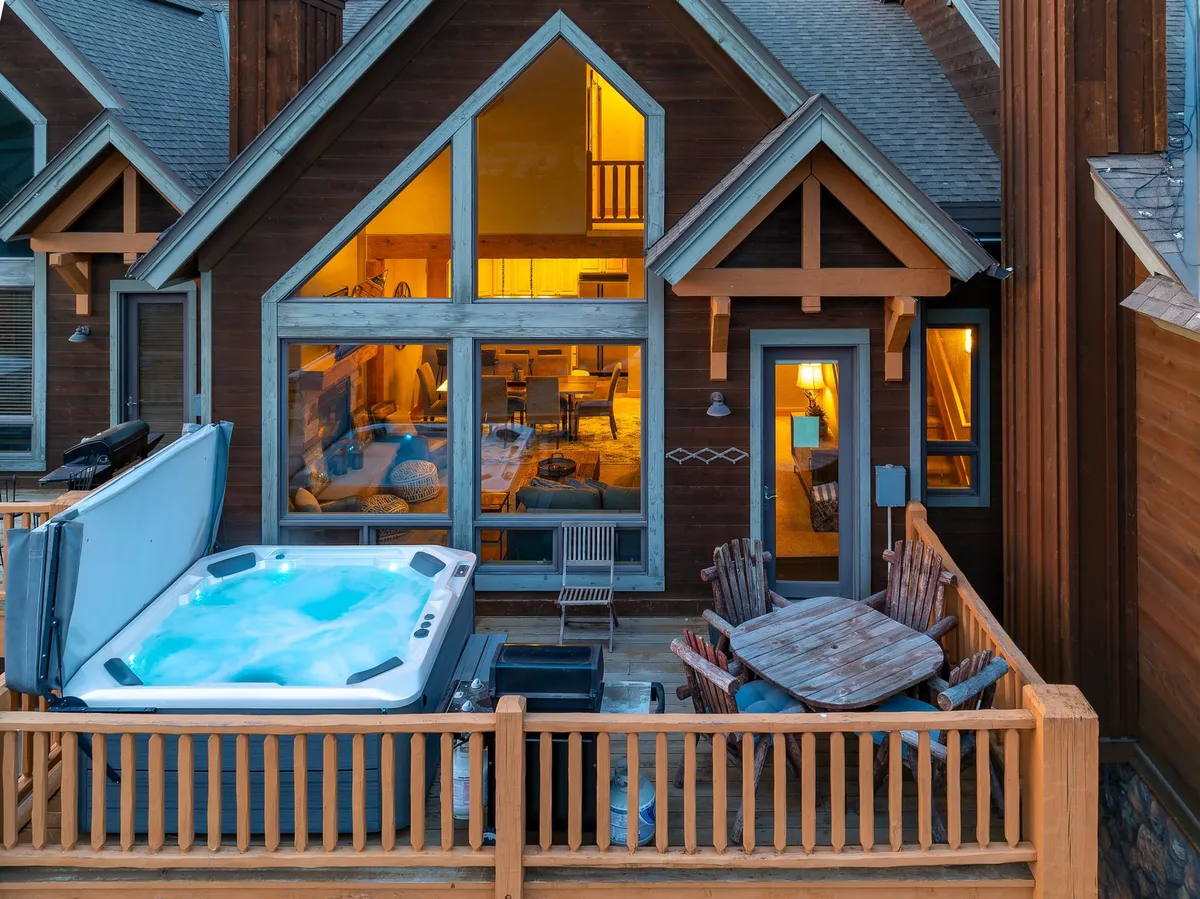 A spacious exterior deck with a jacuzzi, outdoor dining area, wooden deck, and large windows, all set against a warm sunset backdrop.