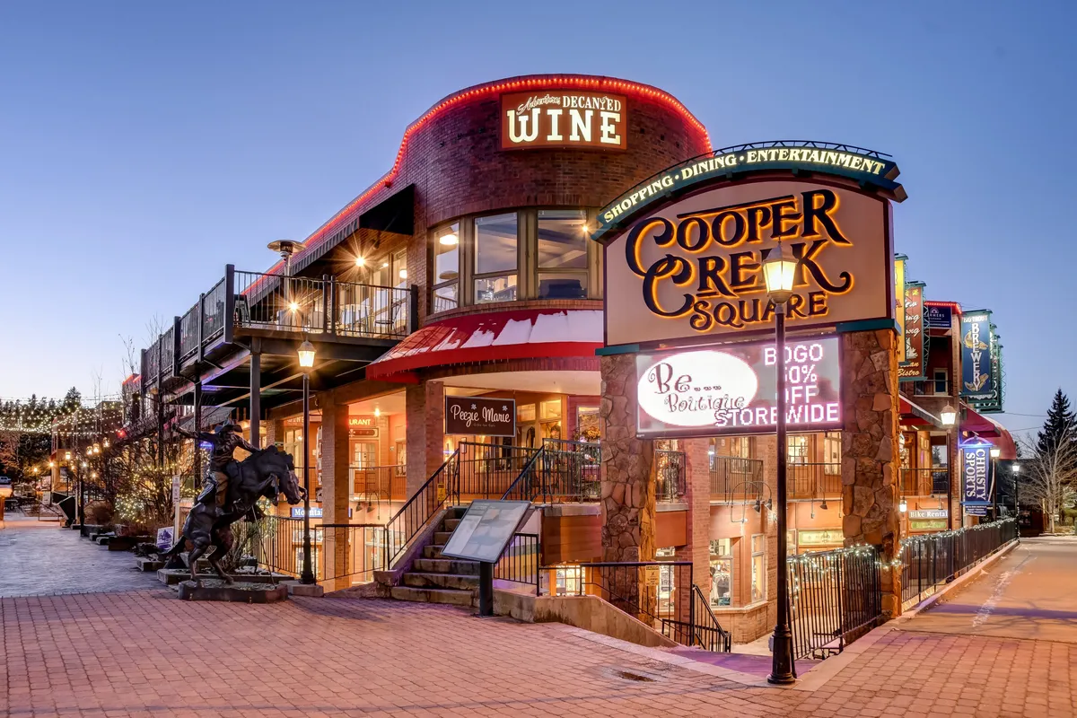 A bustling exterior shot of Cooper Creek Square with its signature sign, decorative lighting, wine shop entrance, and outdoor seating area. The scene is lively, hinting at opportunities for shopping, dining, and entertainment in Winter Park.