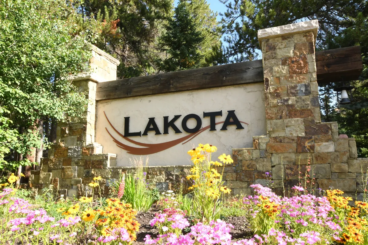 The image captures a stone entrance sign surrounded by vibrant flower beds and lush greenery, all set against the beautiful mountain landscape. This entrance hints at an exclusive and tranquil retreat, welcoming visitors to the mountain experience.